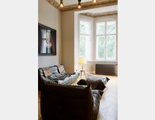 Wohnzimmer mit hohen Decken und dekorativ verziertem Stuck an den Rändern. Der Raum verfügt über große, helle Fenster mit Blick ins Grüne, ein schwarzes Ledersofa mit passenden Hockern, eine Stehlampe sowie ein Bild an der Wand, dazu ein Holzboden im Fischgrätenmuster.