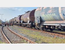 Güterbahnhof mit einer Reihe von Kesselwagen auf den Gleisen. Im Vordergrund sind Schienen und ein Prellbock zu sehen, während die Umgebung von Grasflächen und wenigen Bäumen umgeben ist.