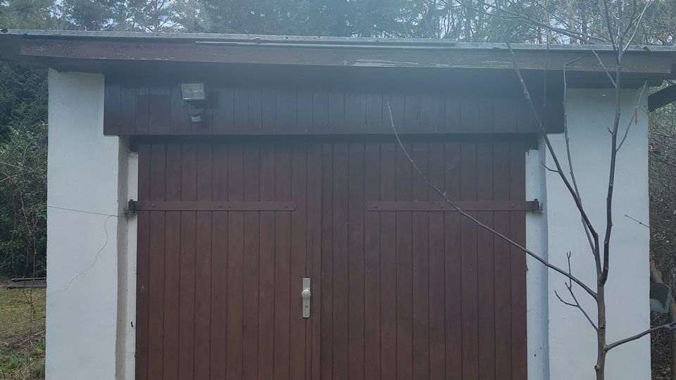 Freistehende, kleine Garage mit weiß verputzten Seitenwänden und großem, zweiflügeligem Holztor in Dunkelbraun unter einem flachen Dachüberstand. Davor unbewachsener Boden mit Grasresten; im Hintergrund Bäume und Sträucher, rechts ein kahler kleiner Baum und ein Verkehrsschild am Rand.