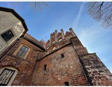 Backsteinkirche aus der Gotik mit Spitzbögen und Ziergiebeln, aufgenommen aus der Froschperspektive. Die Fassade besteht aus rotem Ziegelstein, umgeben von kahlen Bäumen und blauem Himmel.