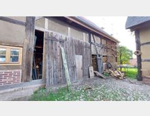 Eine ländliche Scheune mit Fachwerk-Elementen und großen Holztüren. Im Vordergrund liegt gestapeltes Brennholz auf gepflastertem Boden, im Hintergrund sind grüne Bäume und eine Mauer zu sehen.