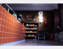 Innenraum einer Bar oder eines Cafés mit einer gemauerten Wand, an der Fußballtrikots und ein eingerahmtes Trikot mit der Nummer "10" hängen. Im Vordergrund befindet sich eine orange geflieste Theke, im Hintergrund ein Regal mit Lebensmitteln und Getränken.