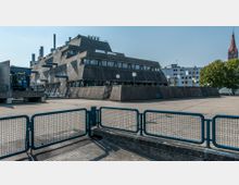 Ein modernes Betongebäude mit pyramidenförmiger Struktur und vielen schrägen Glasfenstern, umgeben von einer gepflasterten Fläche mit blauen Metallzäunen im Vordergrund. Im Hintergrund sind ein Kirchturm, Wohngebäude und Bäume zu sehen.