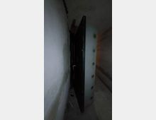 Ein schmaler, spärlich beleuchteter Gang mit Betonwänden, in dem eine schwere Metalltür mit Verriegelungsmechanismen leicht geöffnet ist.