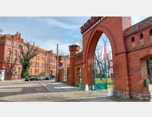 Backsteinbaukomplex mit einem historischen Torbogen aus rotem Ziegelstein, der die Aufschrift "Technische Hochschule Brandenburg" trägt. Im Hintergrund ein mehrstöckiges Gebäude mit gotischen Elementen, flankiert von Bäumen und Kopfsteinpflaster im Vordergrund.