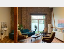 Wohnzimmer mit rustikaler Backsteinwand, großen Fenstern mit Blick auf Bäume, Holzbalken, einem Holzboden und modernen Möbeln, darunter ein grünes Sofa, Sessel und ein Couchtisch. Links ein dekorativer Kamin, rechts ein Lowboard mit Dekoration und Pflanzen.