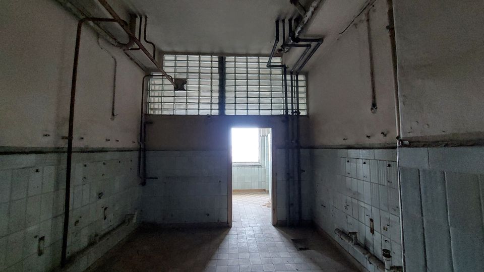 Ein leerer, heruntergekommener Raum mit hellblauen Fliesen an den unteren Wänden und freiliegenden Rohren an der Decke. Im Hintergrund befindet sich eine Tür mit Glasbausteinen darüber, die zu einem helleren Raum führt.