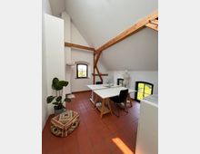 Helles Dachgeschosszimmer mit Schrägdach und sichtbaren Holzträgern, eingerichtet mit einem weißen Schreibtisch, schwarzen Stühlen, einer Schneiderpuppe und einer Pflanze. Der Raum hat Terrakottafliesenboden, zwei Fenster mit Rundbögen und weiße Wände.