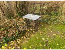 Ein kleiner, rustikaler Gartentisch mit Metallgestell und Schachbrettmuster auf der Tischplatte steht auf einer Wiese, begleitet von einem einfachen Klappstuhl aus Holz. Im Hintergrund sind kahles Gestrüpp, herbstliche Blätter und Efeuranken zu sehen.