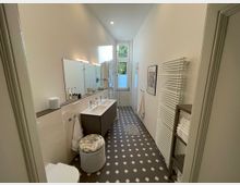 Modernes Badezimmer mit cremefarbenen Wänden, einem dunklen Fliesenboden mit weißem Muster und einem großen Fenster, das Licht hereinlässt. Links befinden sich ein Doppelwaschbecken mit Wandspiegeln und rechts ein Handtuchheizkörper sowie ein Regal mit Handtüchern.