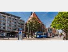 Straßenansicht einer Stadt mit einer historischen Kirche aus Backstein mit spitzem roten Dach im Hintergrund, flankiert von einem modernen Wohn- und Geschäftsgebäude. Im Vordergrund fährt eine blau-weiß gestaltete Straßenbahn auf Schienen, umgeben von Bäumen und Fußgängern.