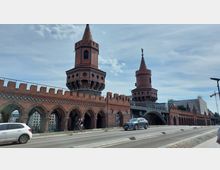 Backsteinbrücke mit zwei Türmen im gotischen Stil, darunter eine Reihe von Rundbögen; auf der Straße davor fahren Autos und ein Fahrradfahrer ist zu sehen. Im Hintergrund sind moderne Gebäude und ein heller Himmel.