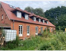 Backsteingebäude mit roten Dachziegeln und mehreren Dachgauben, umgeben von wild bewachsener Vegetation. Mehrere große Fenster und eine Terrasse mit Pflanzen deuten auf einen Wohn- oder Bauernhausstil hin, im Hintergrund sind Bäume zu sehen.