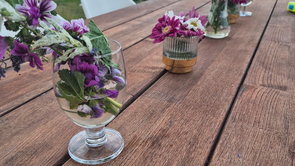 Holz-Gartentisch auf einer Terrasse mit mehreren Glasvasen voller lila- und weißfarbener Blumen; im Hintergrund ein bepflanzter Garten mit Zaun und moderne Wohnhäuser.