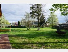 Grüne Wiese mit vereinzelten Bäumen, darunter blühende Obstbäume, in einem umzäunten Gartenbereich. Links steht eine hölzerne Sitzbank mit Überdachung, daneben ein gepflasterter Weg; im Hintergrund sind Gebäude und kleine Schuppen zu sehen.