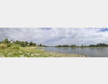 Flusslandschaft mit Kies- und Wiesenufer, im Hintergrund eine kleine Ortschaft mit einer Kirche, umgeben von Bäumen und Wiesen, unter einem bewölkten Himmel.