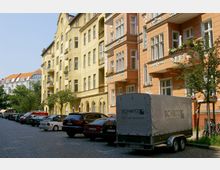 Straßenszene mit Altbau-Wohnhäusern im Berliner Stil, gelb und orange gestrichen, mit dekorativen Fassadenelementen und Balkonen. Im Vordergrund stehen parkende Autos und ein Anhänger mit Plane auf einer gepflasterten Straße, gesäumt von kleinen Bäumen.