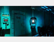 Ein futuristischer Raum mit einem geschlossenen Türportal, das mit „03 Deck“ beschriftet ist, sowie leuchtenden elektronischen Geräten an den Wänden. Der Raum ist in blau-grünem Licht beleuchtet, mit technischen Elementen wie Kabeln, Bildschirmen und Arbeitsplätzen mit Computern.