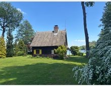 Kleines dunkles Holzhaus mit Reetdach, Schornstein und gelben Fensterläden auf einer großen grünen Wiese, umgeben von hohen Bäumen und blühenden Sträuchern. Im Hintergrund ist ein Seeufer unter blauem Himmel zu sehen.