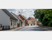 Straßenszene in einer Kleinstadt mit Häusern in verschiedenen Baustilen, darunter weiß verputzte Gebäude, Backsteinhäuser und Fachwerkfassaden, rechts begrenzt von einer Baumreihe. Die Straße ist leicht kurvig, mit Kopfsteinpflaster auf den Gehwegen und Straßenlaternen im klassischen Design.