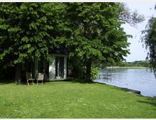 Kleines Holzhaus mit großen Fenstern, umgeben von Bäumen, am Ufer eines Sees mit einem Steg; im Vordergrund eine Wiese mit zwei leeren Holzstühlen.