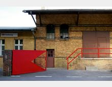 Backsteinfassade eines Gebäudes mit großen Fenstern, einer braunen Tür und geschlossenen Holzläden. Im Vordergrund befinden sich ein rotes Tor, eine Treppe mit rotem Geländer und ein Schild mit der Aufschrift „DB Autozug“.