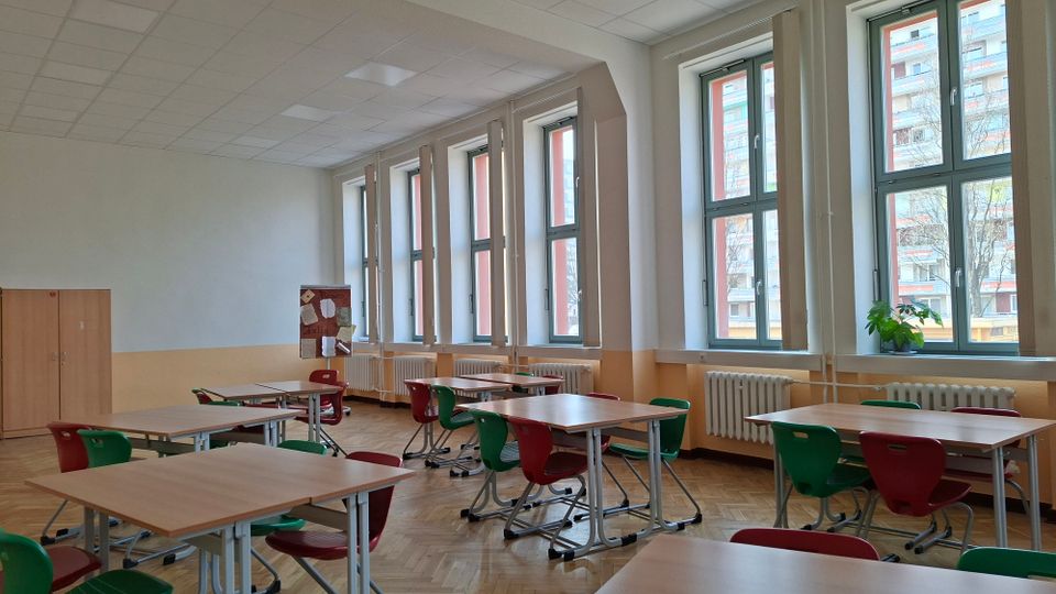 Heller Klassenraum mit mehreren Gruppentischen und rot-grünen Stühlen auf Parkettboden. An einer Wand befinden sich große Fenster mit Heizkörpern darunter, dazu ein Schrank und eine Pinnwand.