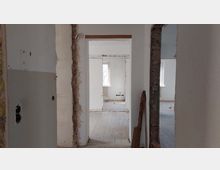 Ein unrenovierter Innenraum mit freiliegenden Ziegelmauern und durchbrochenen Türrahmen. Der Holzboden ist sichtbar, und im Hintergrund befindet sich ein Fenster, durch das Tageslicht einfällt.