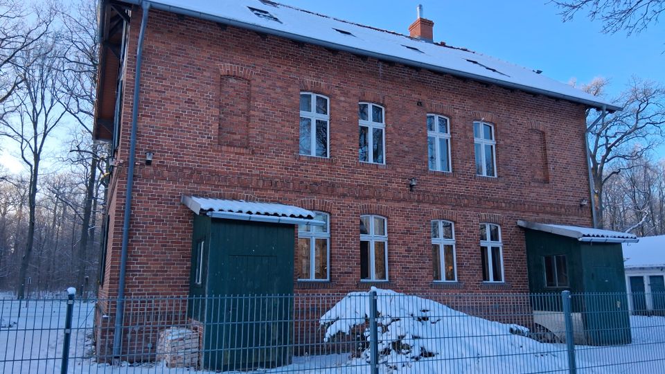 Backsteingebäude mit zwei Stockwerken, symmetrischen Fenstern und schneebedecktem Satteldach, umgeben von einem Zaun und schneebedeckter Landschaft; im Hintergrund sind Bäume eines Waldes zu sehen.