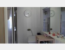 Schmaler Innenraum mit hellen Holzpaneel-Wänden, einer runden Wanduhr und einem großen Wandspiegel über einer Ablage. Daneben stehen ein kleiner Holztisch und ein Hocker; links ist eine Glastür zu einem angrenzenden Raum sichtbar.