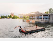 Eine Szene am Wasser mit einem schwimmenden Holzsteg, von dem eine Person ins Wasser springt, während eine andere darauf sitzt. Im Hintergrund sind moderne Gebäude am Ufer, Bäume und zwei Kirchtürme sichtbar.