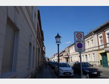 Straßenansicht in einer Wohngegend mit gepflastertem Gehweg, parkenden Autos und historischen Gebäuden mit hellen Fassaden. Ein Laternenpfahl und Verkehrsschilder für eine 30er-Zone und ein Halteverbot sind sichtbar, der Himmel ist klar und blau.