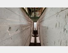 Schmaler Gang in einem industriellen Umfeld, umgeben von hohen, grau gestrichenen Betonwänden mit Kratzspuren und Verschmutzungen. Im Hintergrund sind Metallkonstruktionen und Rohre sichtbar.