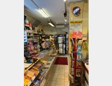 Ein kleiner Kiosk mit Regalen voller Zeitschriften, Süßigkeiten, Getränke und Tabakwaren. Im Vordergrund eine Verkaufstheke, im Hintergrund ein Bereich mit Zeitschriftenständern und Getränkeflaschen.