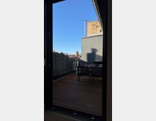 Blick auf eine Terrasse mit Holzboden, einem Tisch und Stühlen, umgeben von Sichtschutzwänden aus Schilfrohr. Im Hintergrund sind Teile von Gebäuden unter klarem, blauem Himmel sichtbar.