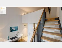 Helle Inneneinrichtung eines modernen Hotelzimmers mit einer hölzernen Treppe, metallenen Geländern und weißen Wänden. Links steht ein Schreibtisch mit einer Lampe und Dekoration, im Hintergrund ein Türdurchgang zu einem weiteren Raum.