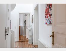 Ein heller Flur mit weiß gestrichenen Wänden und Holzfußboden; im Vordergrund eine weiße Statue sowie ein Heizkörper an der Wand, weiter hinten eine geöffnete Tür, durch die ein weiterer Raum mit einer Büste auf einem Sockel sichtbar ist. An den Wänden hängen farbige Kunstwerke.