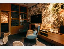 Gemütlicher Lounge- oder Wohnzimmerbereich mit gemusterter Tapete, zwei gepolsterten Sesseln und einem wandmontierten Fernseher über einem Holzsideboard. Rechts stehen ein Plattenspieler und weitere Audiogeräte, links trennt eine Holzschiebetür mit Gitterelementen den Raum von einer angrenzenden Nische.