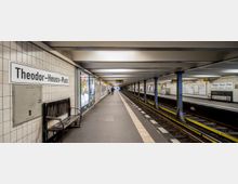 U-Bahn-Station "Theodor-Heuss-Platz" mit zwei parallel verlaufenden Bahnsteigen, die durch Stützpfeiler getrennt sind. Die Wände sind mit hellen Fliesen verkleidet, und es sind Sitzbänke, Werbetafeln sowie Stationsschilder sichtbar; das Gleis verläuft gerade in die Ferne.