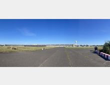 Panoramafoto eines ehemaligen Flughafengeländes mit einer weitläufigen, asphaltierten Fläche im Vordergrund und Wiesen zu beiden Seiten. Im Hintergrund steht das langgestreckte Hauptgebäude mit grauer Fassade, daneben ein markanter Turm mit einer Radarkuppel.