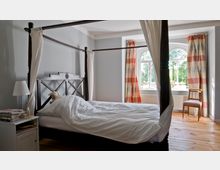 Schlafzimmer mit Himmelbett aus dunklem Holz, weißer Bettwäsche und einem Nachttisch mit Büchern und Lampe. Im Hintergrund ein großes, bogenförmiges Fenster mit gestreiften Vorhängen, das Tageslicht hereinlässt, Holzfußboden und ein einfacher, hölzerner Stuhl.