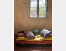 Ein einfacher Raum mit einer cremefarbenen, verwitterten Wand und einem einzelnen Fenster, durch das Tageslicht einfällt und einen Blick auf Pflanzen und eine rote Ziegelmauer bietet. Im Vordergrund liegt ein niedriger Stapel von farbigen Matratzen und verschiedenen dekorativen Kissen.