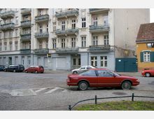 Straßenszene mit einem historischen Wohngebäude mit verzierten Fassaden und Balkonen im Hintergrund. Im Vordergrund sind mehrere parkende Autos auf einer Kopfsteinpflasterstraße zu sehen, rechts davon steht ein kleineres, gelbes Haus mit grünen Fensterläden.