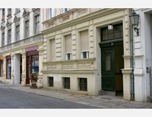 Straßenszene mit historischen Wohngebäuden; links befinden sich Schaufenster eines Ladens mit dekorativen Elementen, rechts ein Eingang mit dunkelgrüner Tür und Hausnummer 4, flankiert von verzierten Fassaden und einem Wandlaternenhalter.