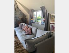 Gemütliches Wohnzimmer mit einer großen grauen Couch, dekoriert mit gemusterten Kissen. Im Hintergrund befindet sich ein Fenster mit grauen Vorhängen, durch das Tageslicht einfällt, sowie Pflanzen und Bilder an den Wänden.