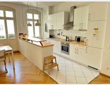 Helle, moderne Wohnküche mit weißen Schränken, Holzarbeitsplatten und einer langen Kücheninsel mit schmaler Ablage. Große Fenster und drei Hängelampen beleuchten den Raum; sichtbar sind Herd mit Edelstahl-Dunstabzug, Spüle sowie ein Mix aus Fliesenboden im Küchenbereich und Fischgrätparkett im Essbereich.