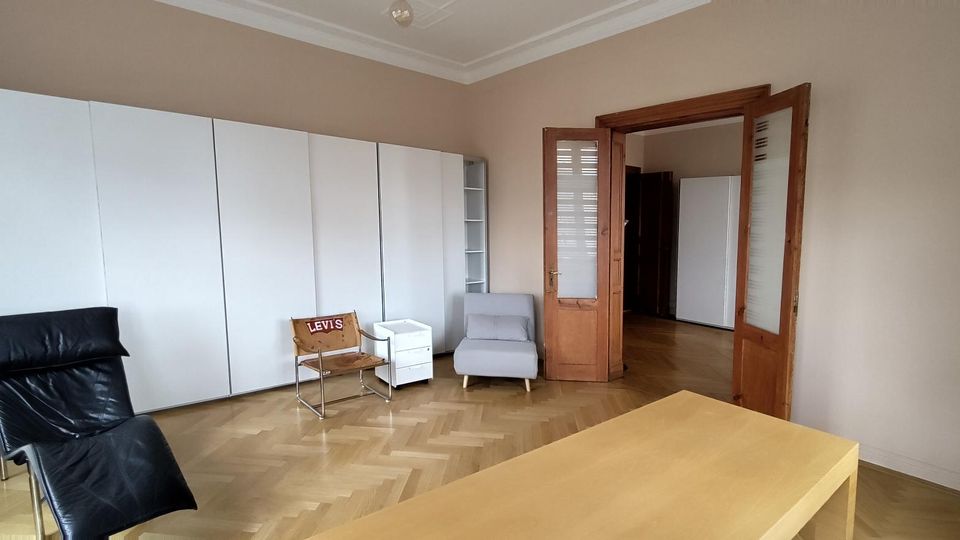 Heller Wohnraum mit Fischgrätparkett, hohen Decken und großem weißen Schrank an der Wand. Offene Holztüren mit Glasfeldern führen in einen angrenzenden Raum; davor stehen ein schwarzer Liegestuhl, ein kleiner Sessel und ein einfacher Tisch.