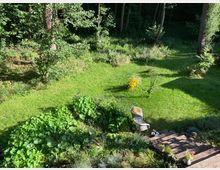 Garten mit gepflegtem Rasen, umgeben von Bäumen und dichter Vegetation. Im Vordergrund eine Holzterrasse mit Pflanzen in Töpfen, ein einzelner Gartenstuhl, und eine gelb blühende Pflanze im Zentrum der Wiese.