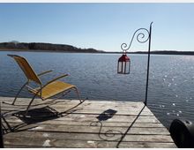 Holzsteg an einem ruhigen See mit einem gelben Schaukelstuhl und einer roten Laterne an einem Metallhaken. Im Hintergrund ist eine Baumreihe am Ufer unter klarem, blauem Himmel zu sehen.