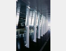 Moderner Innenraum mit einer Reihe von vertikalen, gläsernen Trennwänden, die durch weiße Metallhalterungen befestigt sind. Die Decke besteht aus perforierten Metallplatten, und der Boden ist dunkel und reflektierend.